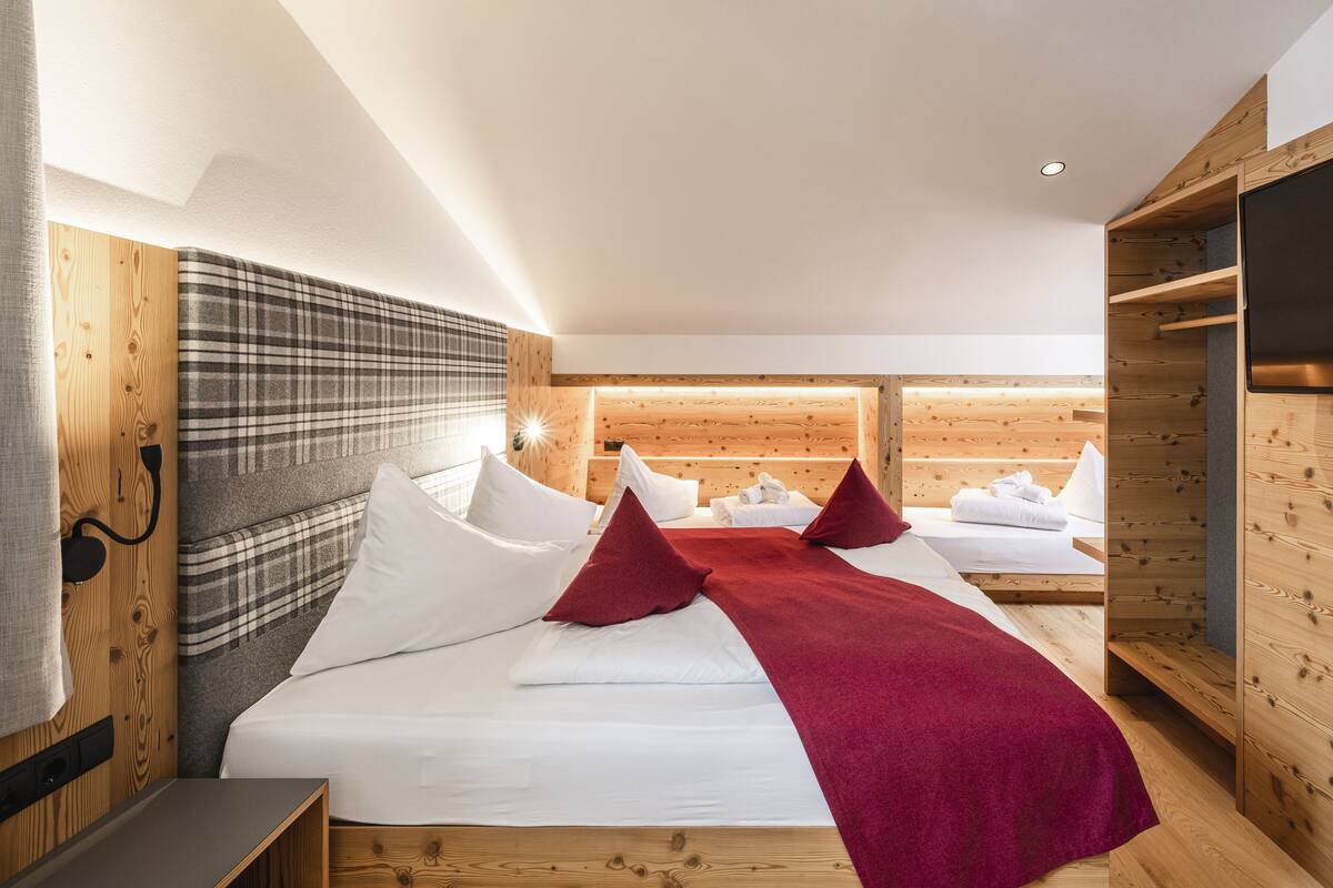 A modern hotel room with a wooden headboard, a large bed with white linens and red pillows, a wooden wardrobe, and a flat-screen TV.