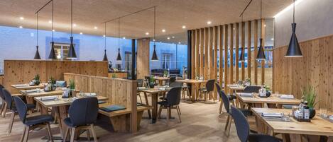 C’est un restaurant moderne, doté de mobilier en bois et d’un plafond haut. Les tables sont disposées pour accueillir les clients, et de grandes fenêtres offrent une vue sur l’extérieur.