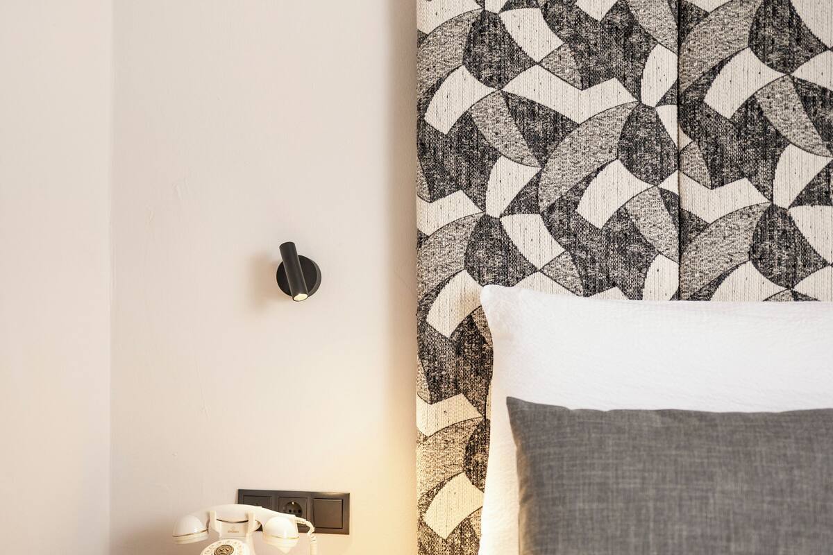 A neatly made bed with a patterned headboard, a bedside table with a telephone, and a wall-mounted light fixture.