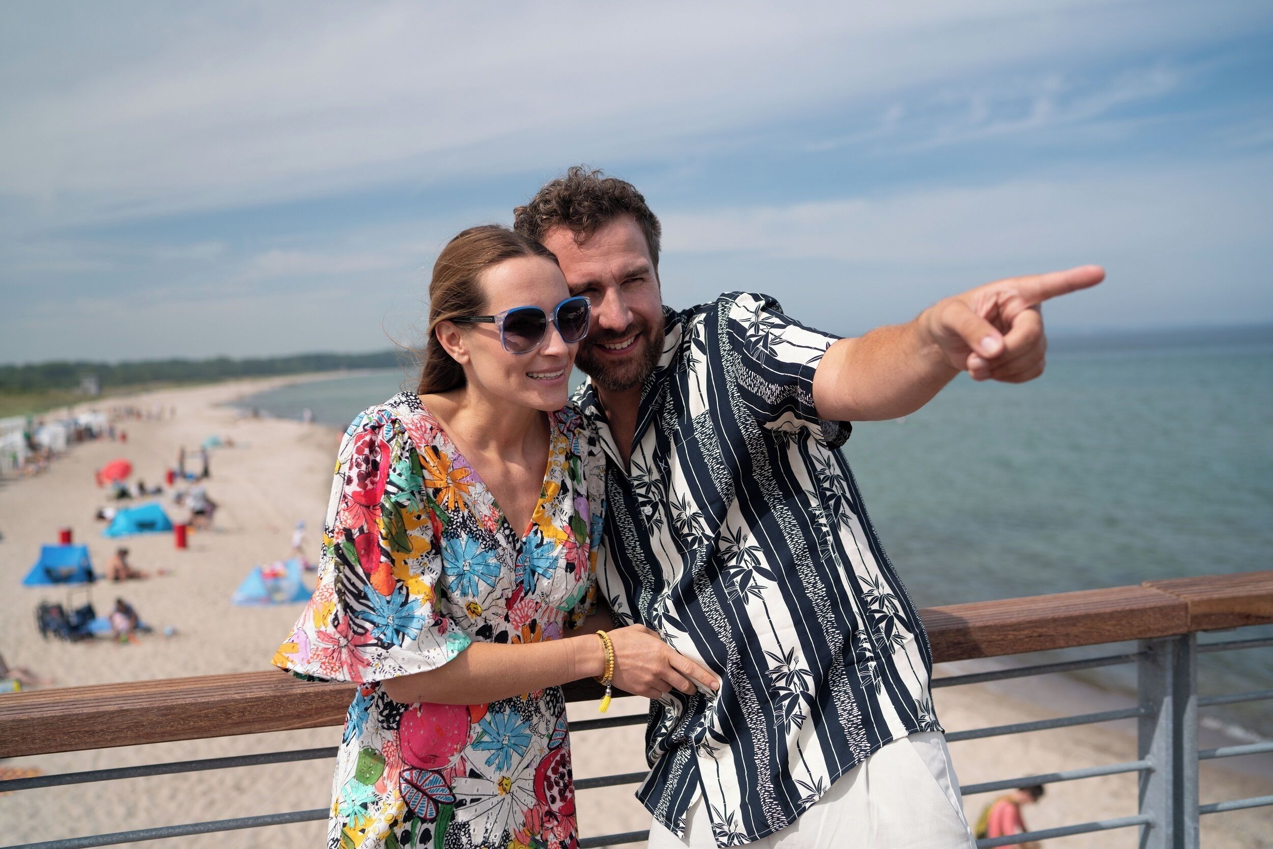 A couple on a beach with a railing, pointing towards the sea.