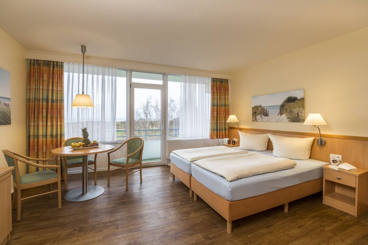 Une chambre d’hôtel avec un lit, deux tables de chevet, une chaise, une petite table avec des fruits et une fenêtre avec des rideaux.