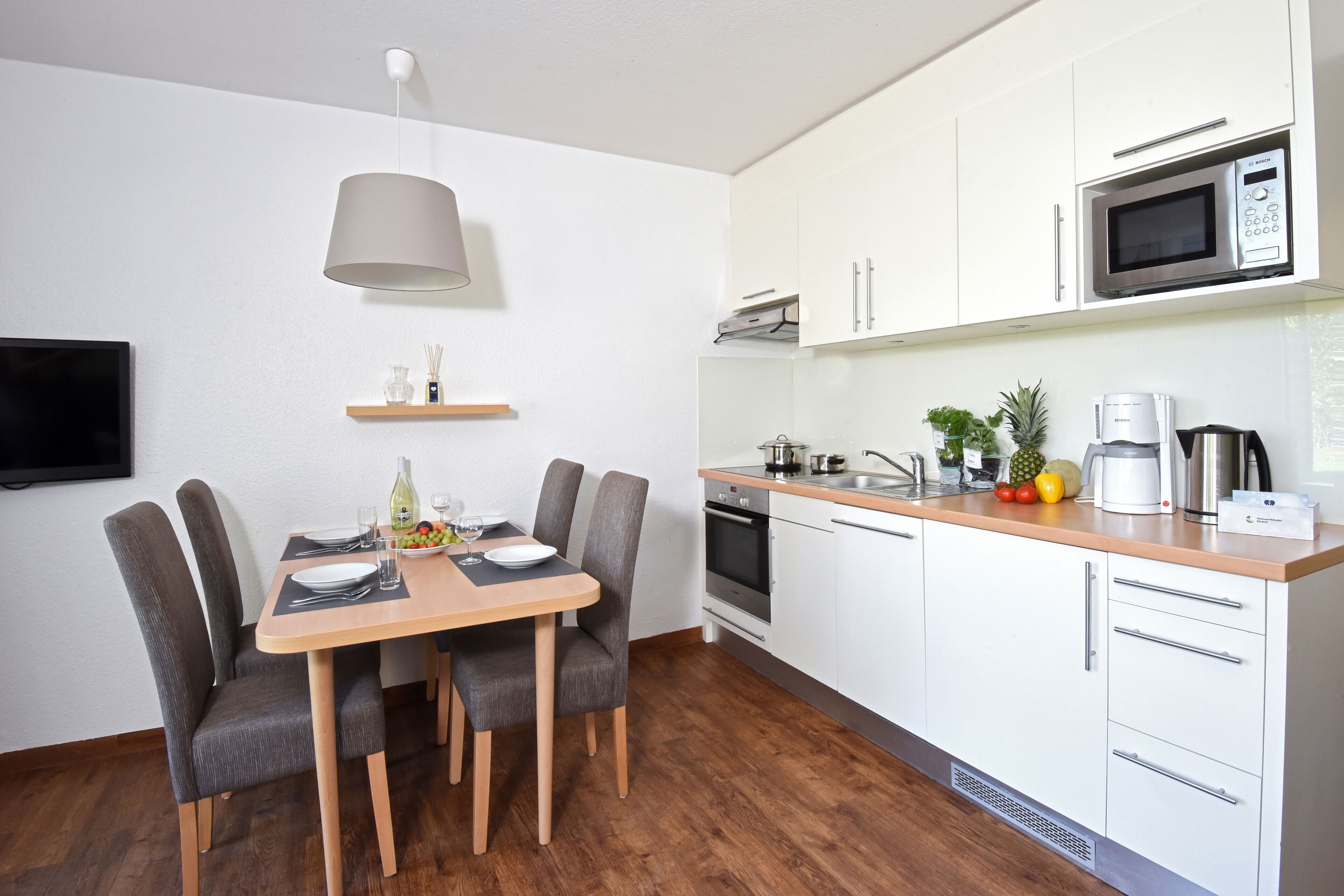 Une cuisine moderne avec des meubles blancs, un four à micro-ondes et une machine à café. On y trouve une table à manger avec des chaises, ainsi qu’un téléviseur fixé au mur.
