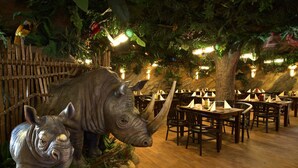 Un restaurante con mesas y sillas de madera, una gran estatua de rinoceronte y una decoración temática de selva.