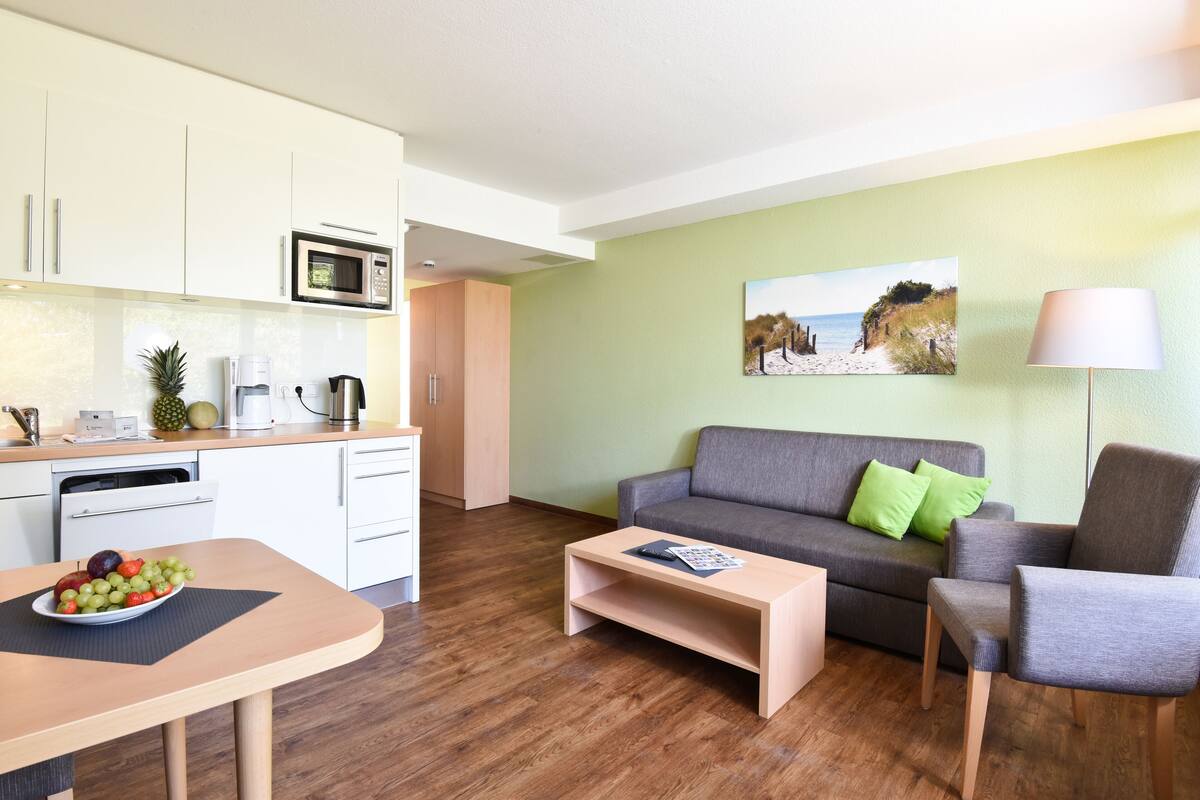 Un salon moderne avec un canapé gris, une table basse et une table à manger agrémentée d’un présentoir de fruits. On y trouve également un coin cuisine avec des meubles blancs et un four à micro-ondes.