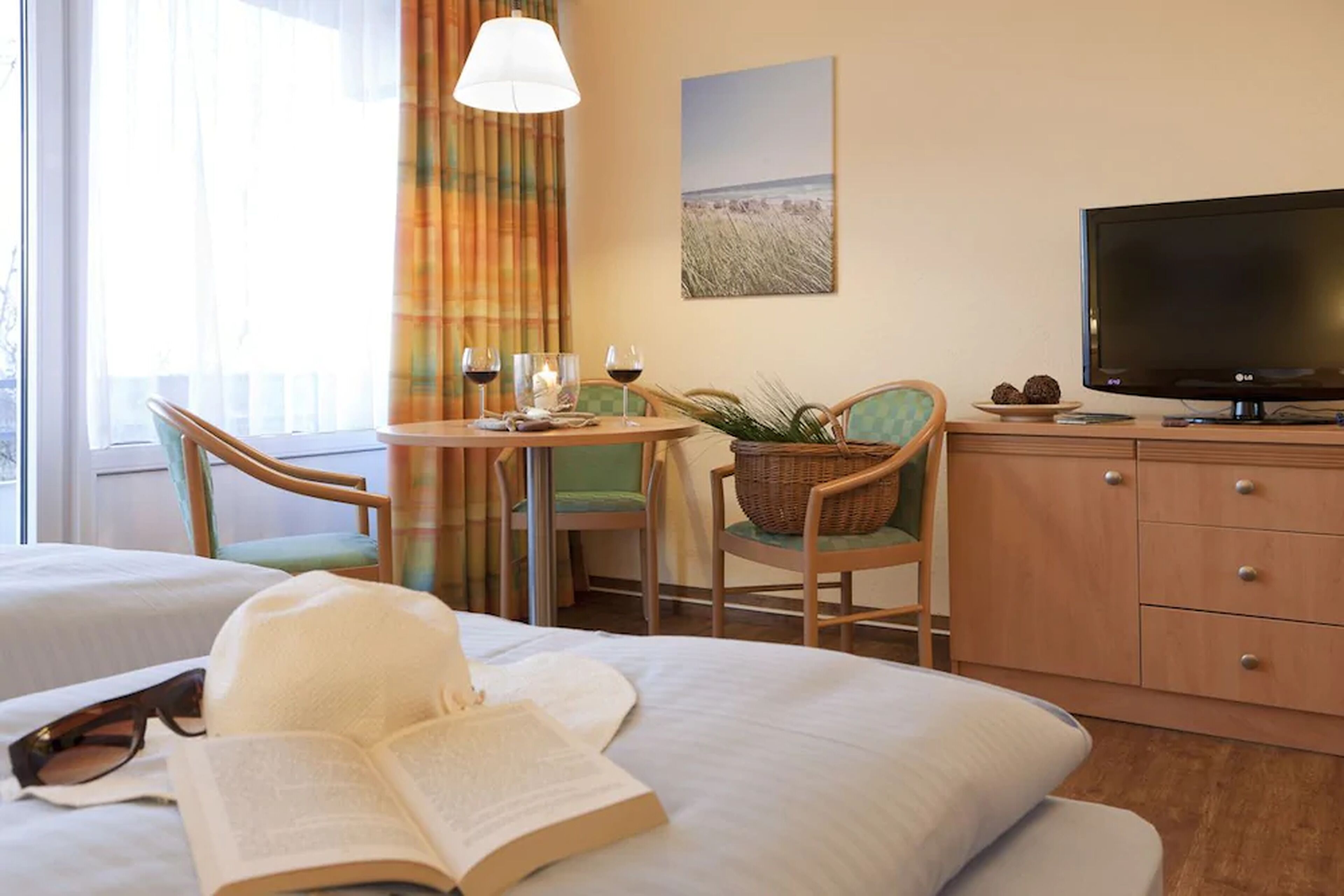 A hotel room with a bed, a reading area with a book, a television, a dining table with chairs, and a view of the beach through the window.
