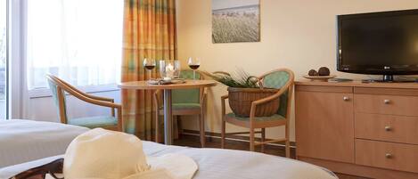 A hotel room with a bed, a reading area with a book, a television, a dining table with chairs, and a view of the beach through the window.