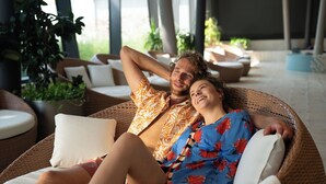 Una pareja descansando en sillones de mimbre en un espacio interior iluminado por el sol, con plantas en macetas y asientos acolchados.