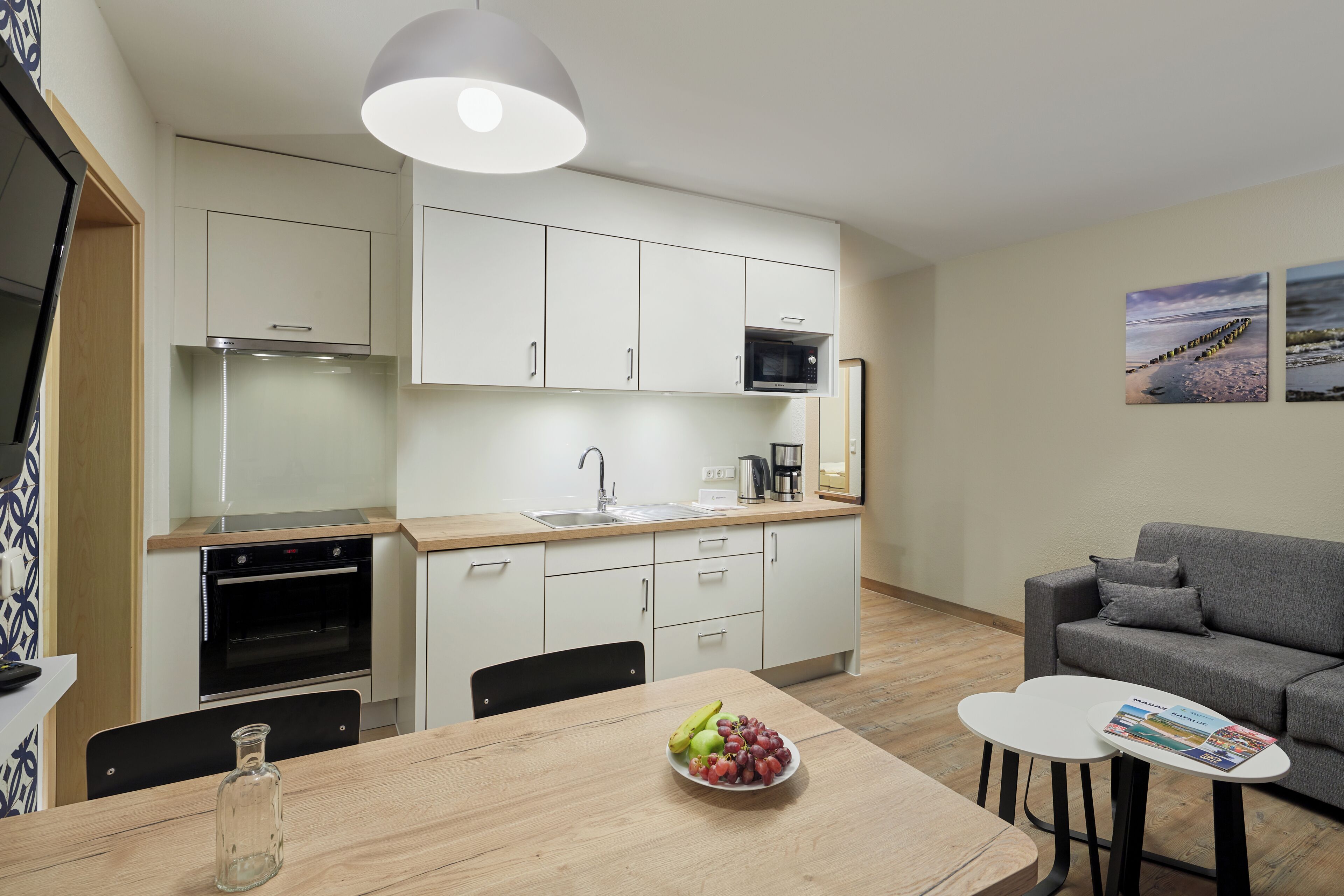 A modern kitchen with white cabinets, a microwave, and a sink. There is a dining table with a bowl of fruit and a magazine. A gray sofa is in the living area.