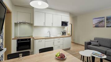 A modern kitchen with white cabinets, a microwave, and a sink. There is a dining table with a bowl of fruit and a magazine. A gray sofa is in the living area.