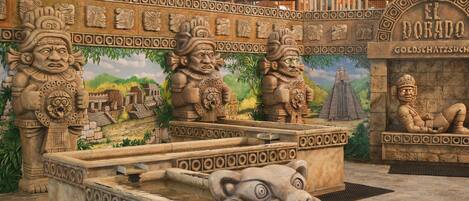 Ein Steinkneippbrunnen mit Löwenkopf, umgeben von Steinmetzarbeiten und einem Wandgemälde eines Maya-Tempels.