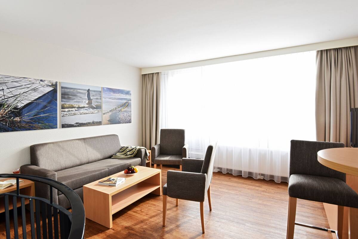 Un salon moderne comprenant un canapé, deux fauteuils, une table basse et une table à manger. Trois tableaux encadrés ornent le mur, et une grande fenêtre est agrémentée de rideaux.