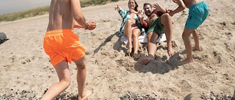 En gruppe mennesker på en sandstrand, hvor nogle sidder og andre står, og beskæftiger sig med aktiviteter ved stranden.
