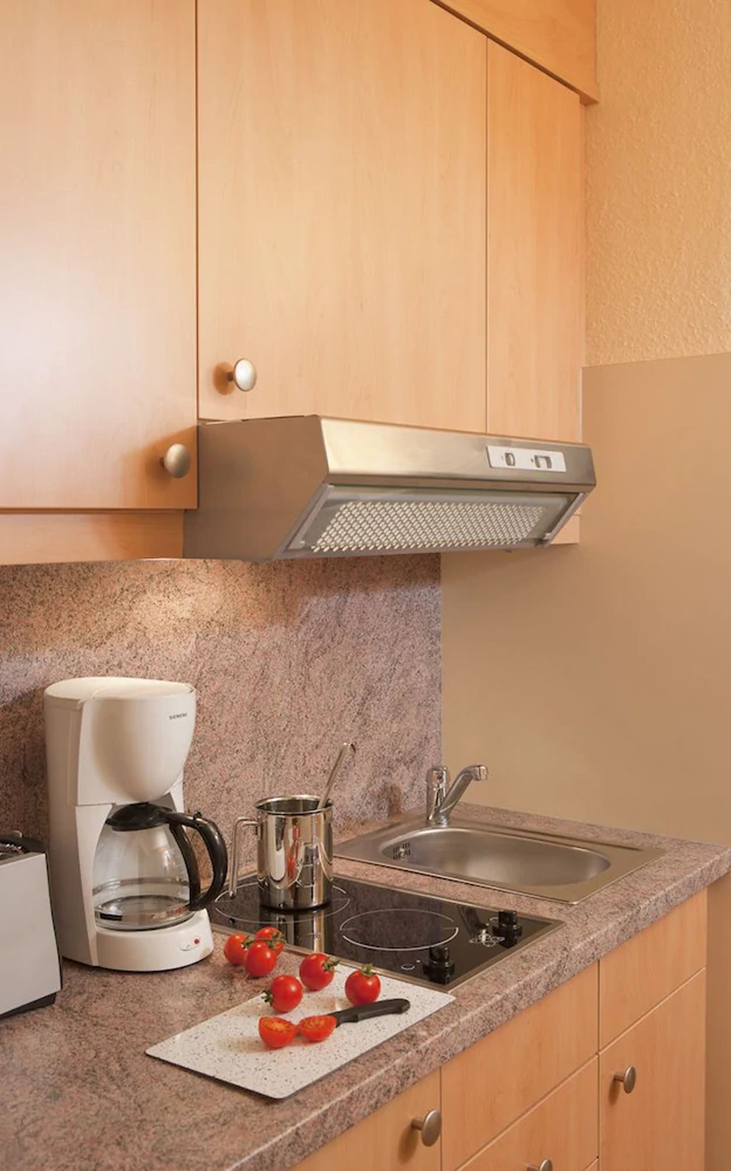 Una cocina con armarios de madera, una campana extractora de acero inoxidable, una cafetera, un fregadero y una tabla de cortar con tomates.