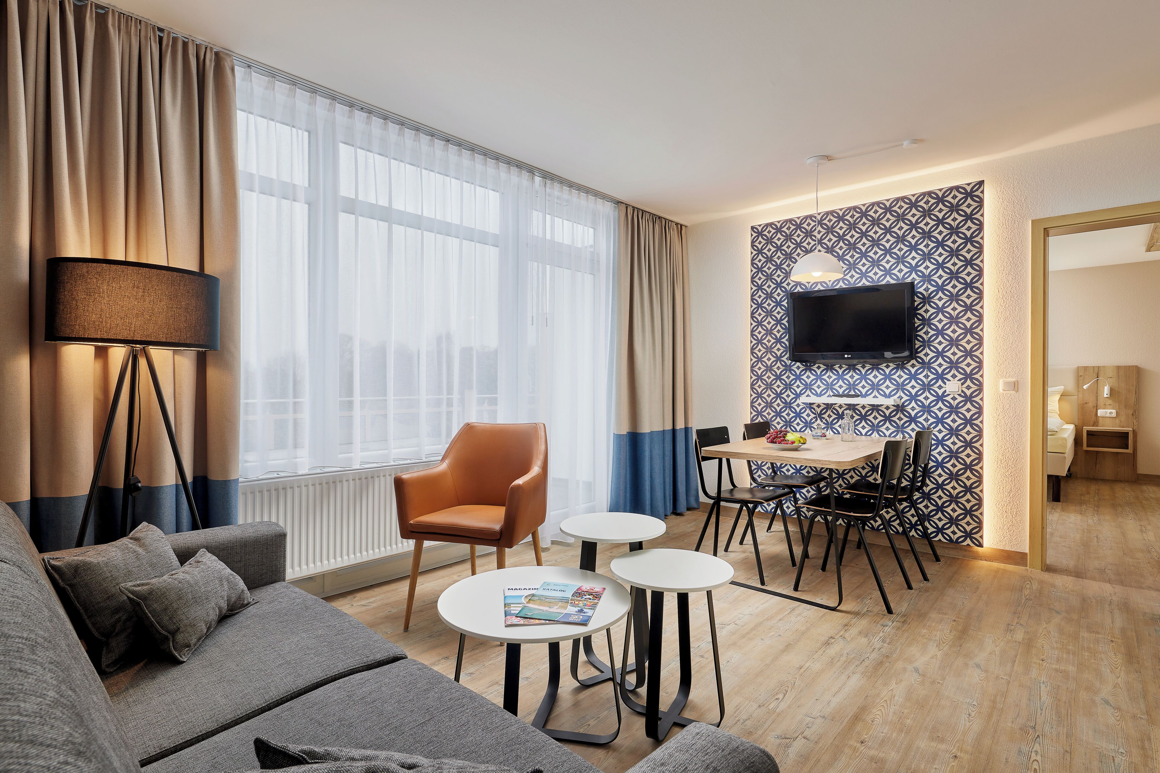 Une chambre d’hôtel moderne, dotée d’un canapé, de deux petites tables, d’un coin repas avec des chaises, d’un téléviseur fixé à un mur orné de motifs et d’une grande fenêtre avec des rideaux.