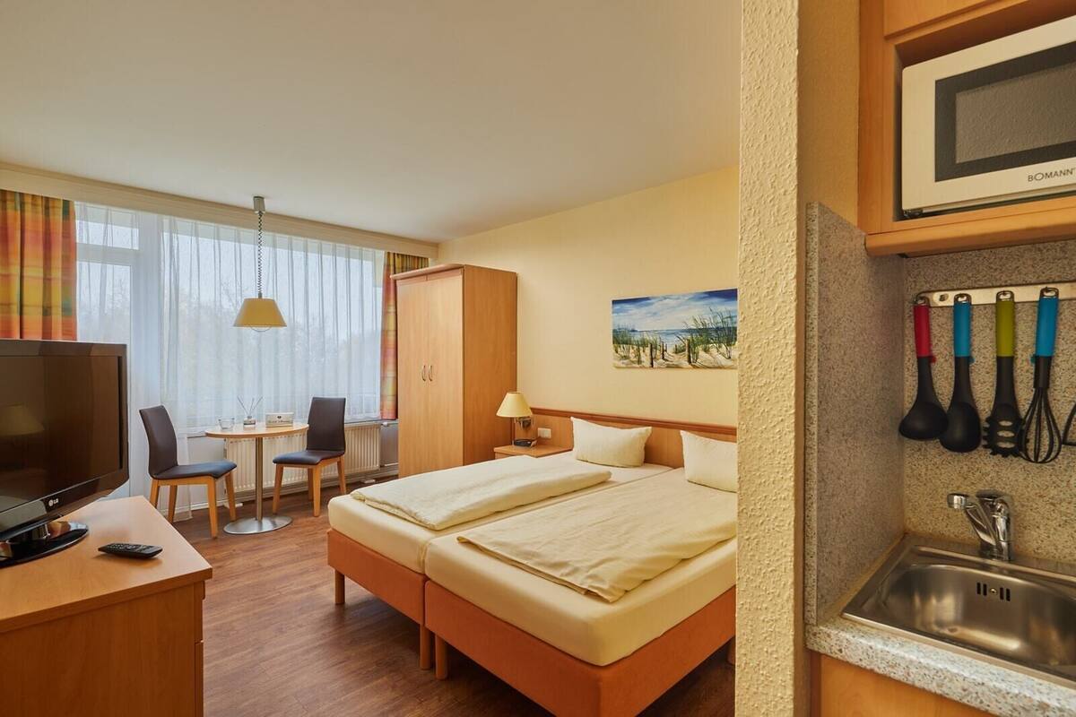 Une chambre d’hôtel comprenant un lit, un bureau avec une télévision, une chaise, une petite table, une fenêtre avec des rideaux, un tableau au mur et un coin cuisine avec un évier et des ustensiles.