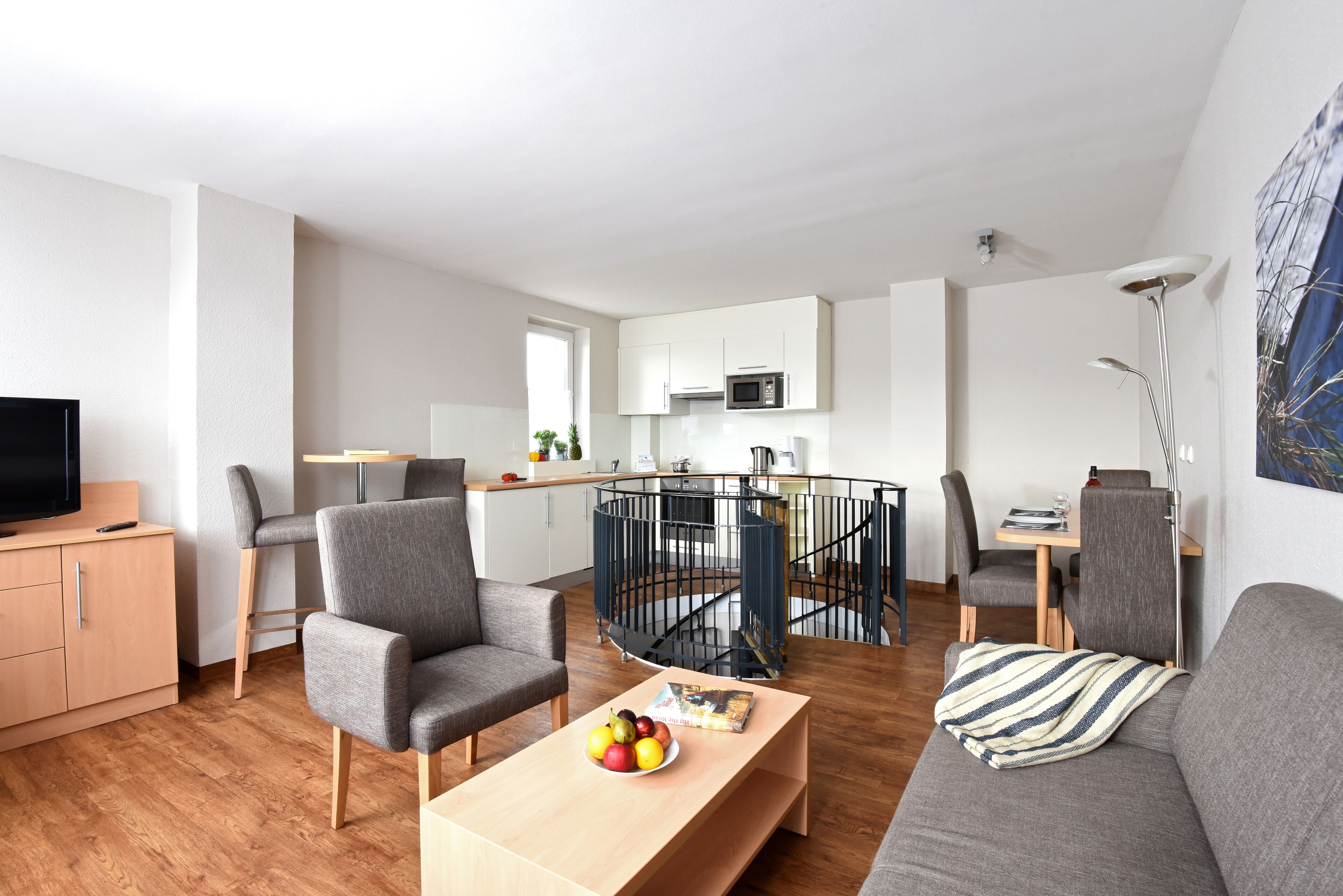 A modern living room with a sofa, armchairs, a coffee table with fruit, a TV, and a kitchen area with a microwave.