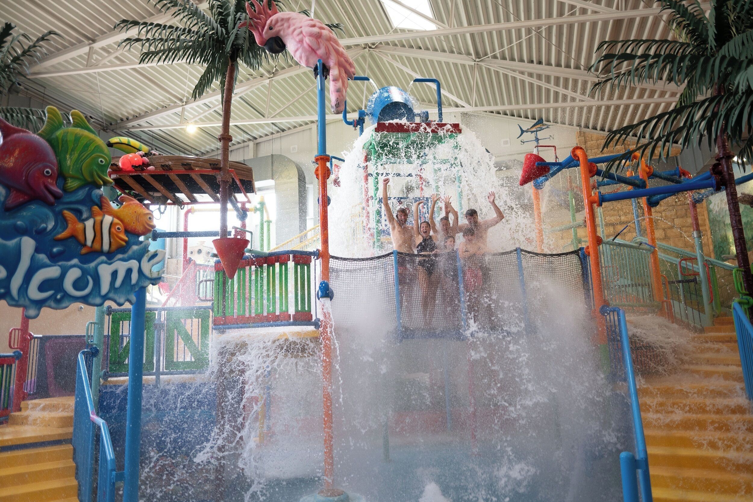 An indoor water play area with slides and water jets, surrounded by colorful decorations and a 'Welcome' sign.