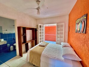 A hotel room with a bed, a ceiling fan, a bathroom with a sink and toilet, and an open door leading to a red outdoor area. - Pousada Pantai Maresias (São Sebastião)