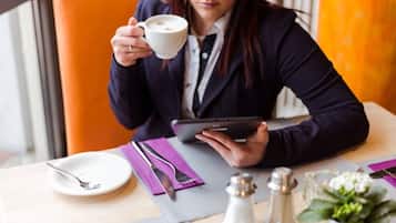A person sitting at a table with a cup of coffee, a tablet, and a plate with cutlery.