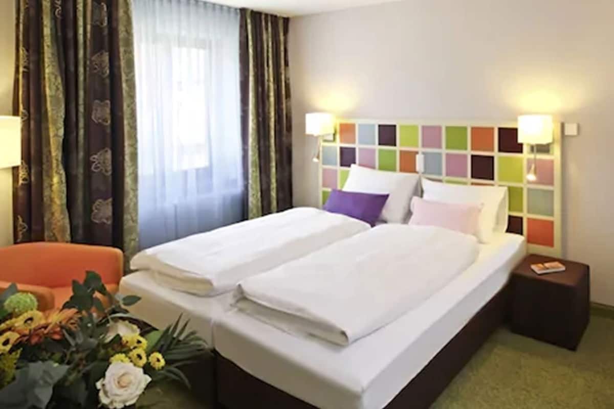 A hotel room with a double bed, a bedside table, a lamp, a chair, and a window with curtains.