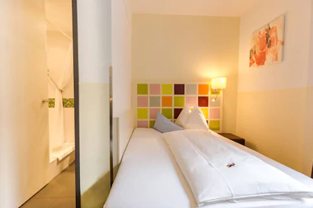 A hotel room with a bed, a colorful headboard, a nightstand, and a wall-mounted lamp.