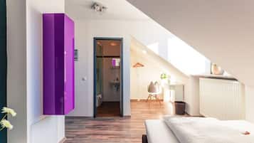 Une chambre moderne avec un plancher en bois, un lit recouvert d’une couverture blanche, une armoire violette et une vue sur la salle de bain par une porte ouverte.