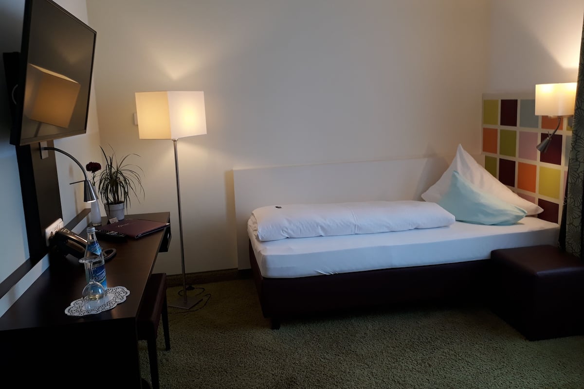 A hotel room with a bed, a desk with a lamp, a television, and a colorful wall panel.