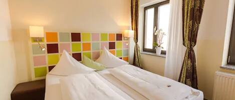 A hotel room with two beds, a colorful headboard, a window with curtains, and a radiator.