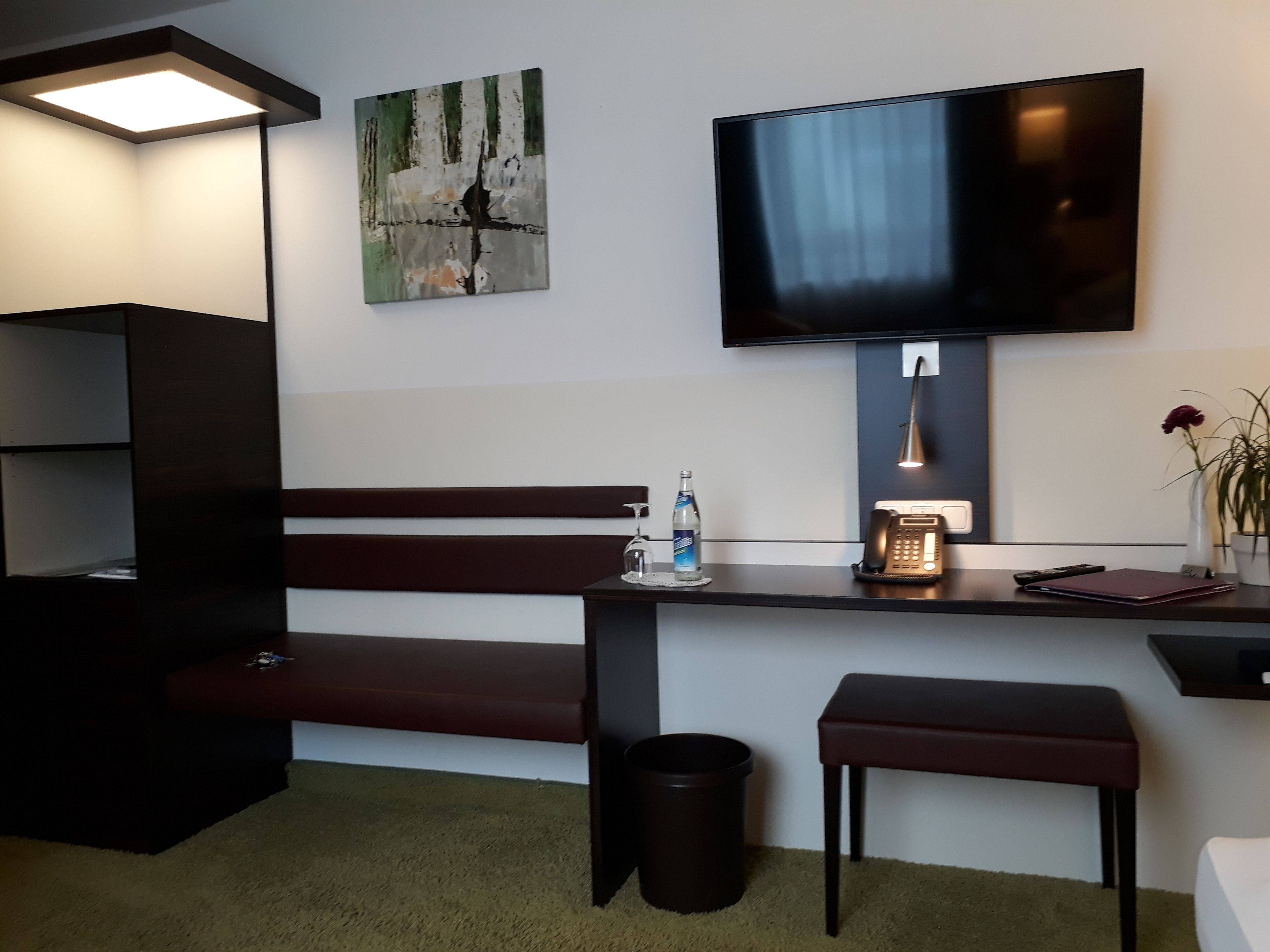 A modern hotel room with a dark wooden bench, a small table with a bottle and a phone, a trash can, and a TV mounted on the wall.