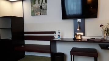 A modern hotel room with a dark wooden bench, a small table with a bottle and a phone, a trash can, and a TV mounted on the wall.