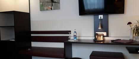 A modern hotel room with a dark wooden bench, a small table with a bottle and a phone, a trash can, and a TV mounted on the wall.