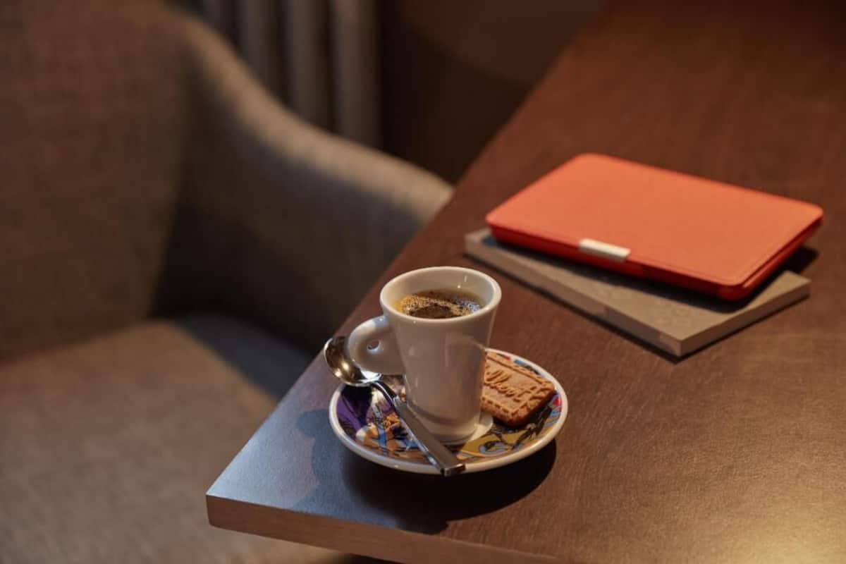 Una taza de café sobre un plato, una galleta, una cuchara y una libreta roja sobre una mesa de madera.