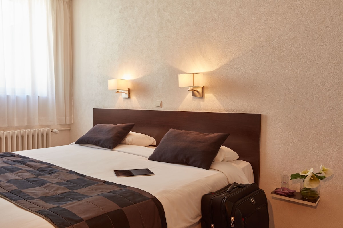 Habitación de hotel con una cama, dos lámparas de pared, una mesita de noche con un jarrón de flores y una maleta.