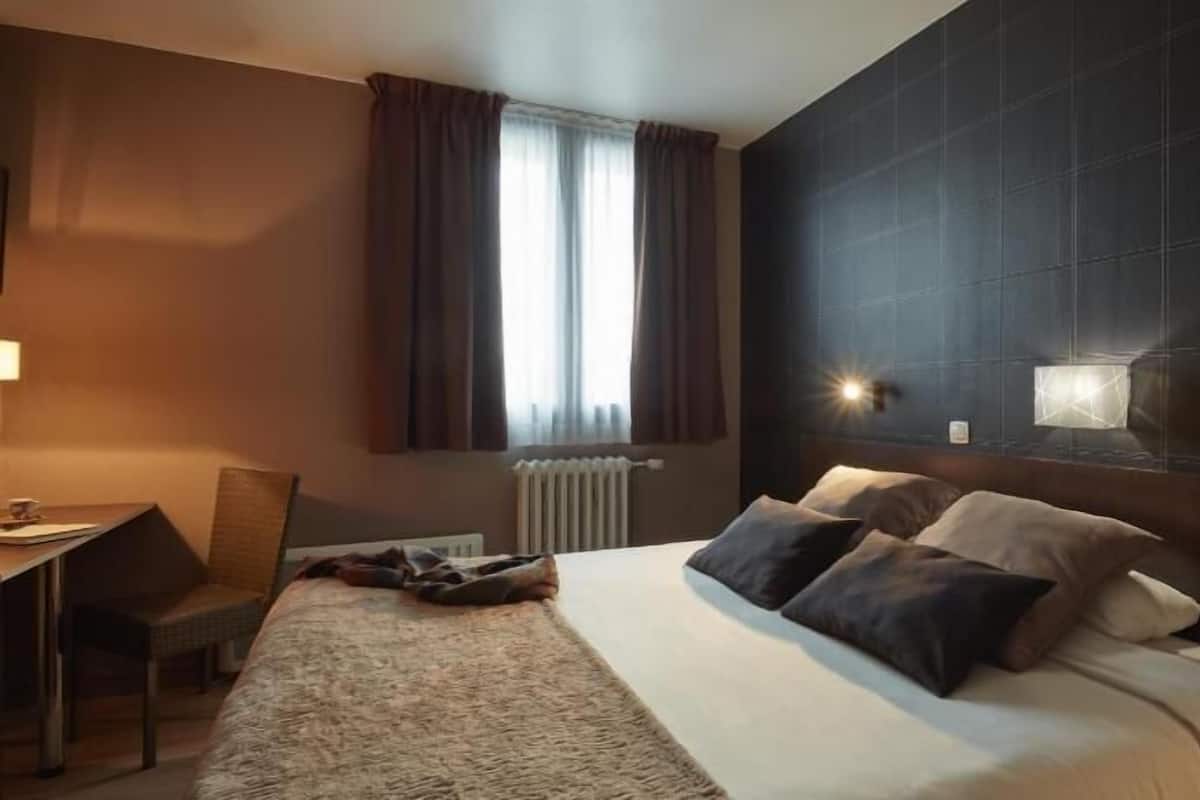 Habitación de hotel con cama, escritorio, silla, ventana con cortinas y lámpara de pared.