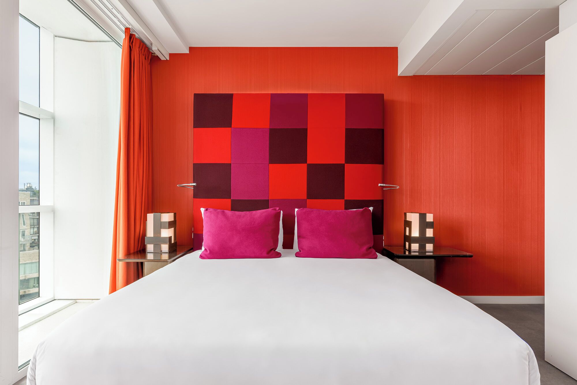 Una habitación de hotel moderna con una cama grande, dos mesitas de noche, un cabecero rojo y negro a cuadros y vistas de la ciudad a través de la ventana.