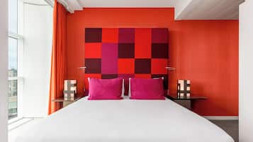 Una habitación de hotel moderna con una cama grande, dos mesitas de noche, un cabecero rojo y negro a cuadros y vistas de la ciudad a través de la ventana.