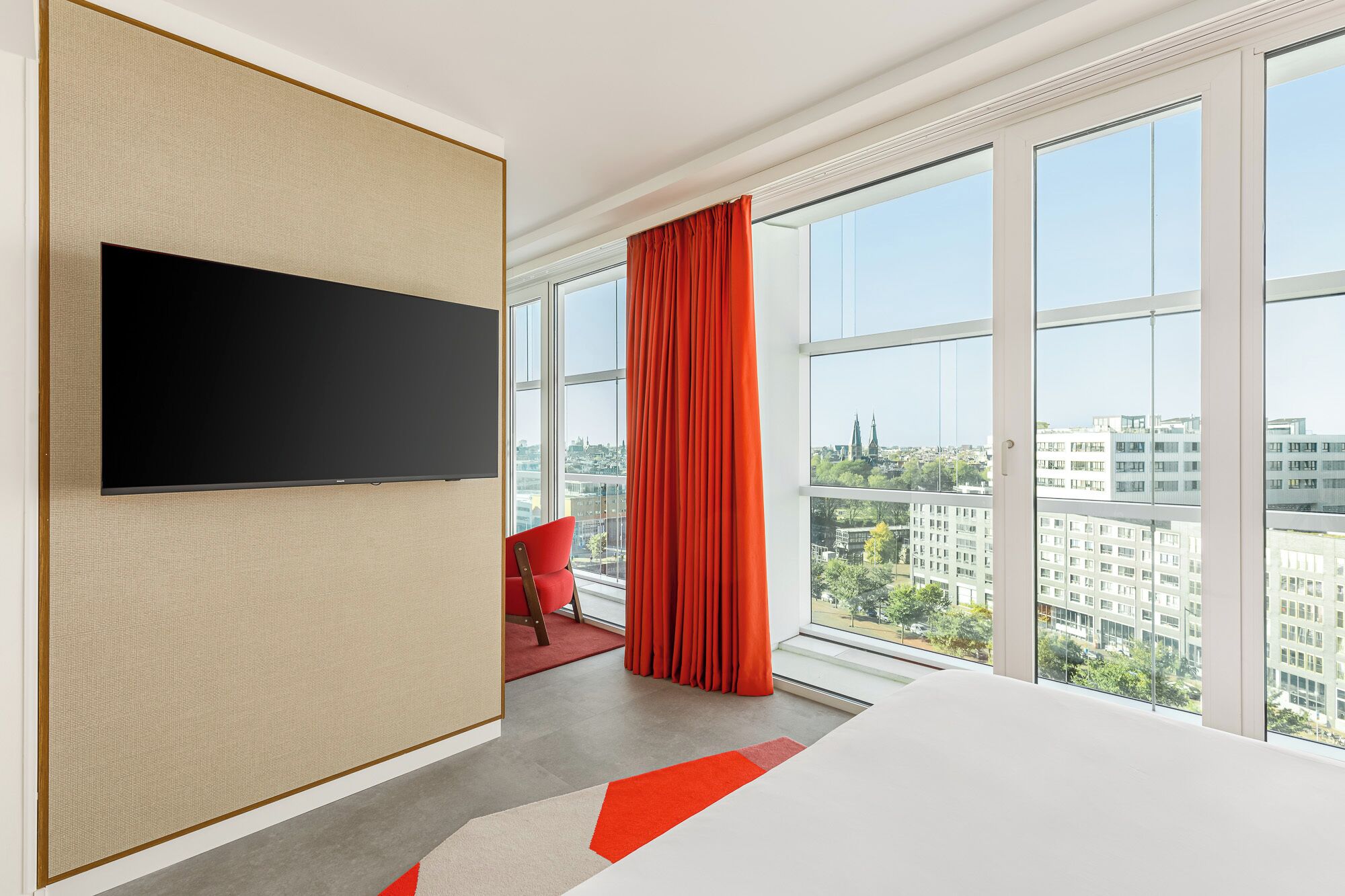 Una habitación de hotel moderna con una cama grande, un televisor de pantalla plana montado en un panel texturizado, una silla roja y ventanas del suelo al techo que ofrecen vistas a la ciudad.