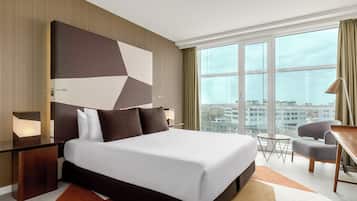 Habitación de hotel con una cama grande, un escritorio, una silla y vistas a la ciudad a través de amplios ventanales.