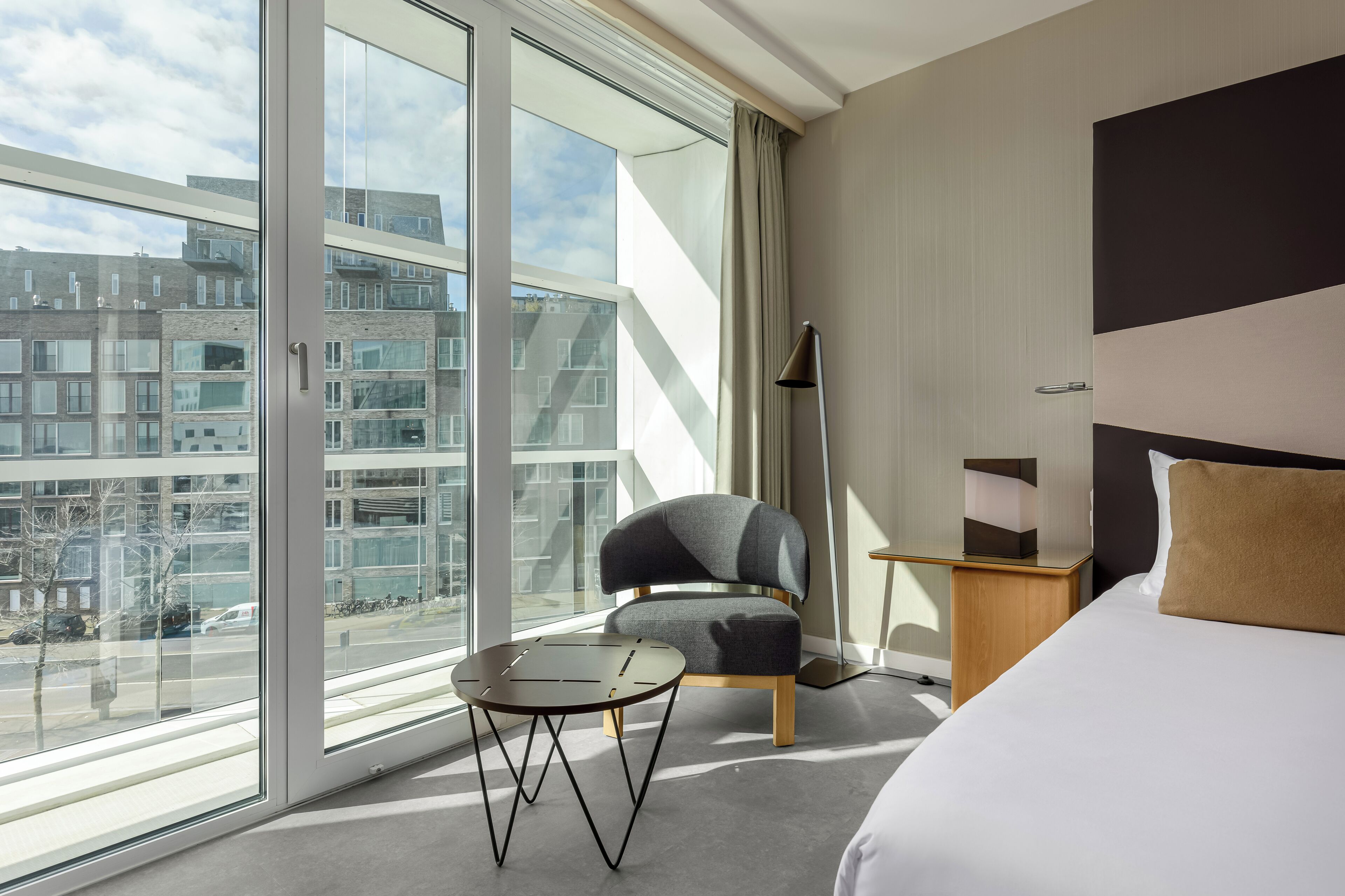 Una habitación de hotel moderna con un ventanal grande, una mesa redonda, un sillón gris, una lámpara de noche y una cama con un cojín marrón.