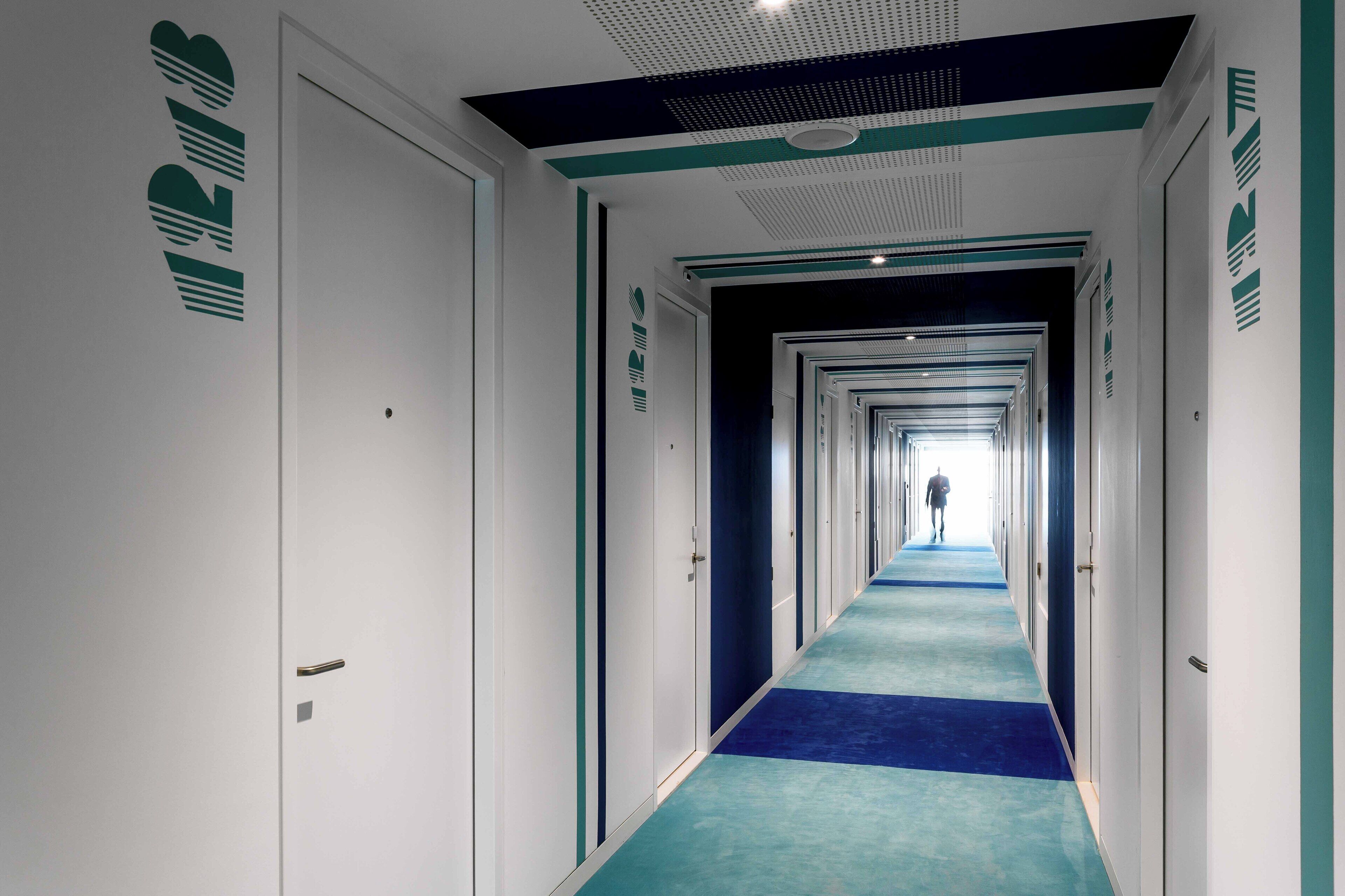 Un largo y estrecho pasillo de hotel con alfombra azul, paredes blancas y puertas a ambos lados.
