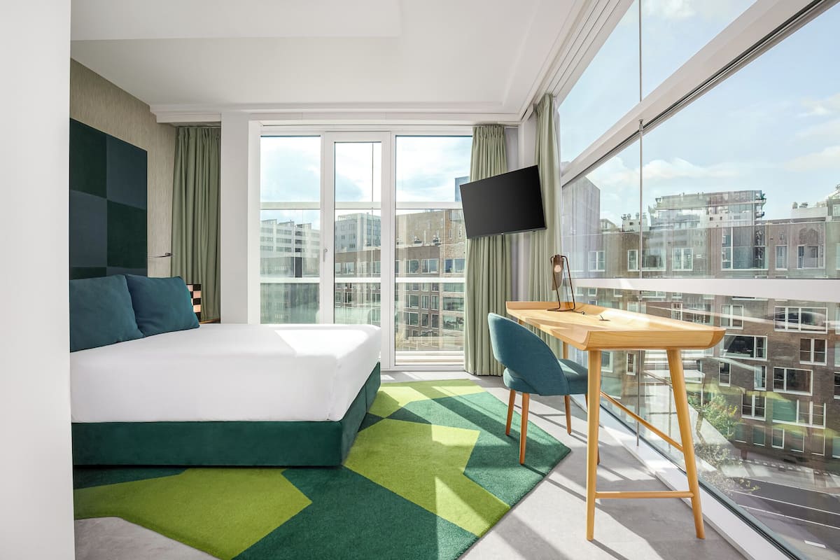 Una habitación de hotel moderna con cama, escritorio y un amplio ventanal con vistas a la ciudad.