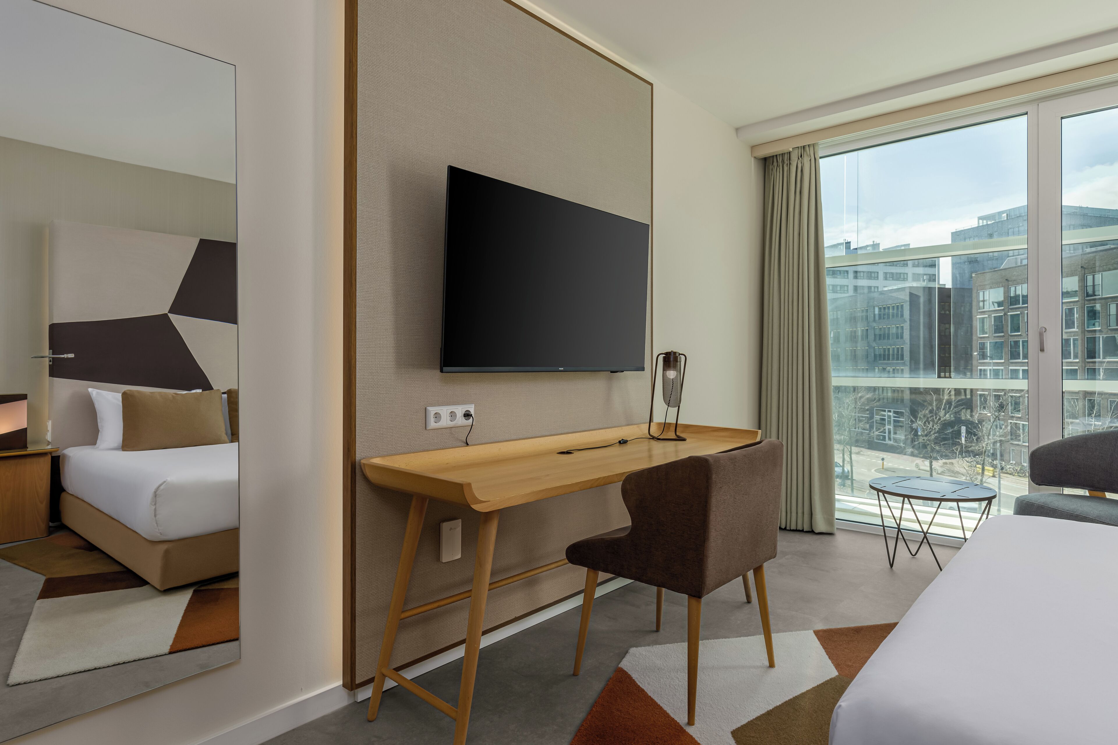 A modern hotel room with a bed, a desk, a chair, a TV, and a balcony with a city view.