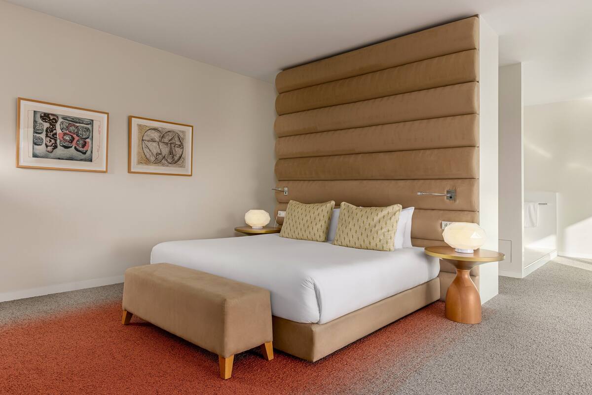 Un dormitorio moderno con una cama grande, un banco, dos mesitas de noche con lámparas y dos obras de arte enmarcadas en la pared.