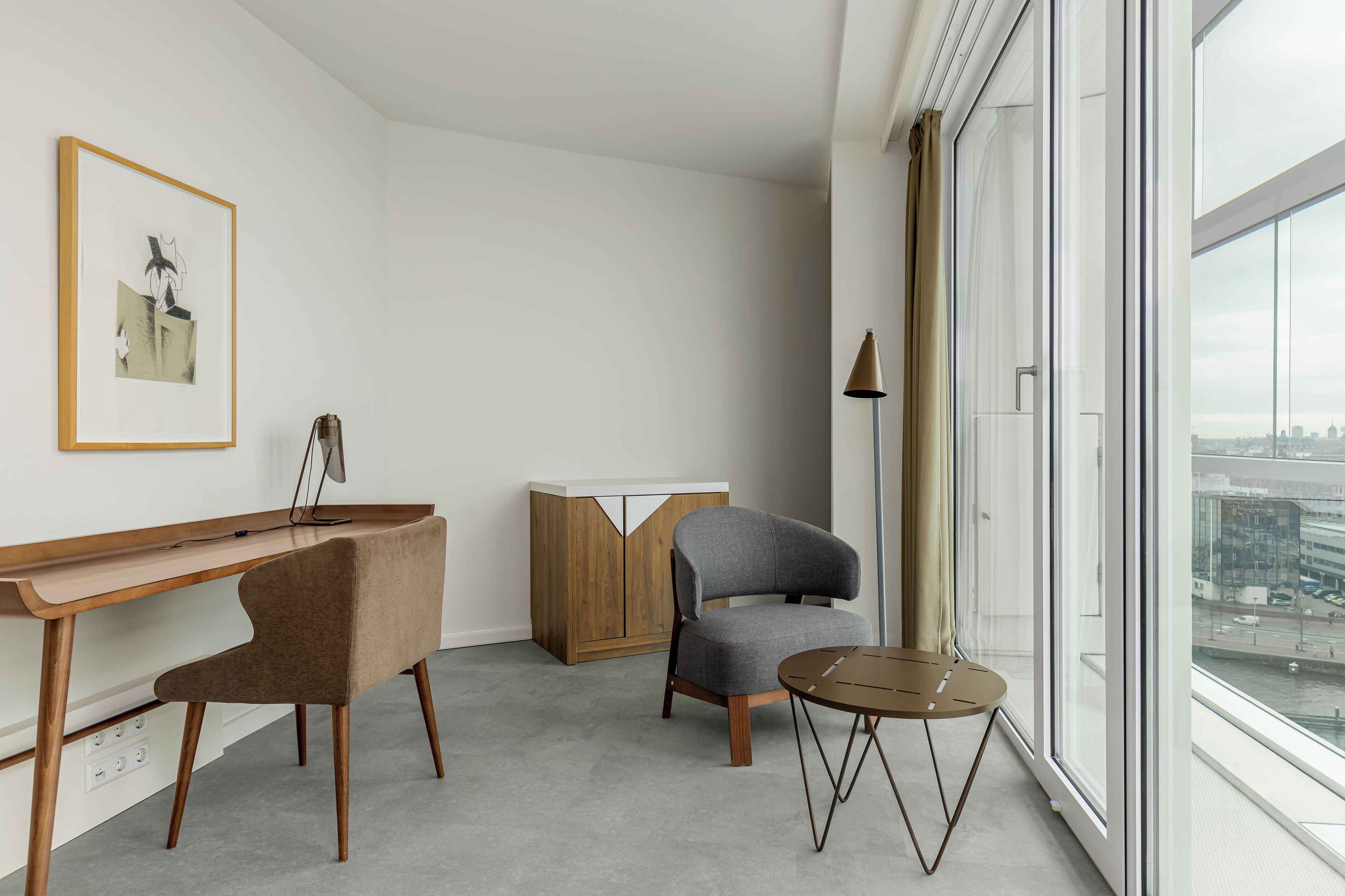 Una habitación moderna con un escritorio de madera, dos sillas, una mesita redonda, un armario y un amplio ventanal con vista a la ciudad.