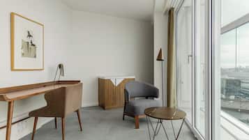 Una habitación moderna con un escritorio de madera, dos sillas, una mesita redonda, un armario y un amplio ventanal con vista a la ciudad.