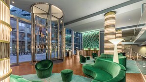 A modern lobby with a glass elevator, green seating area, and a reception desk.