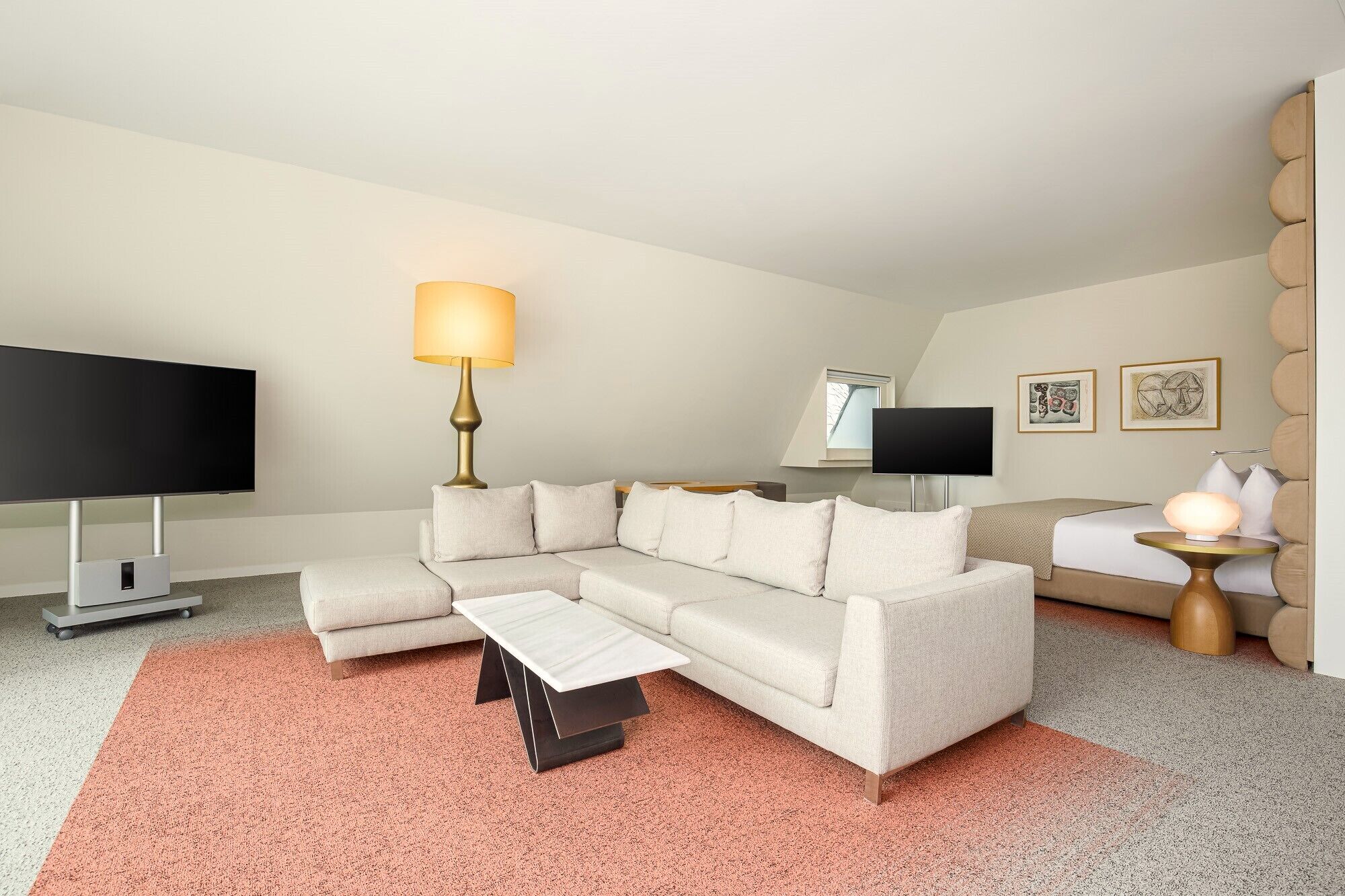 Une chambre d’hôtel moderne dotée d’un canapé d’angle, d’un téléviseur à écran plat, d’un bureau avec une lampe et d’œuvres d’art accrochées aux murs.