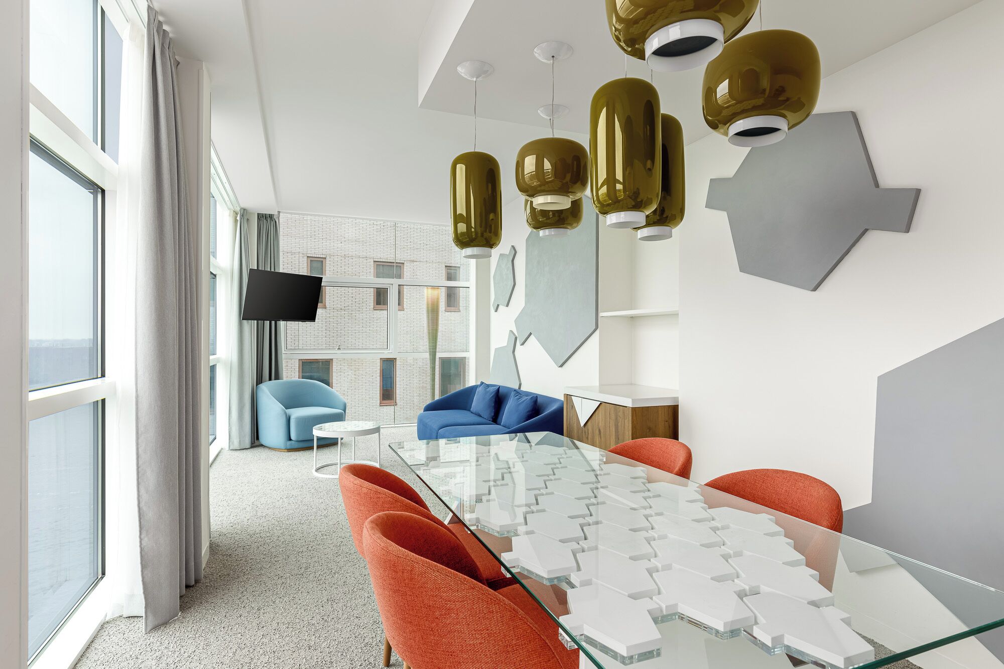 Een moderne hotelkamer met een glazen tafel, oranje stoelen, een blauwe bank en een groot raam.