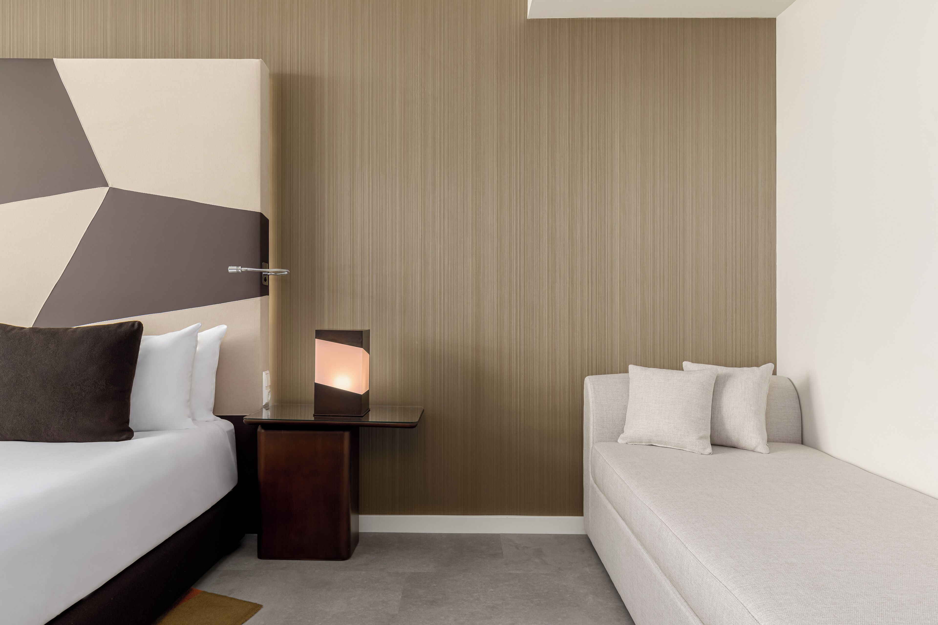 A modern hotel room with a bed, a nightstand, a bedside table with a lamp, and a sofa.