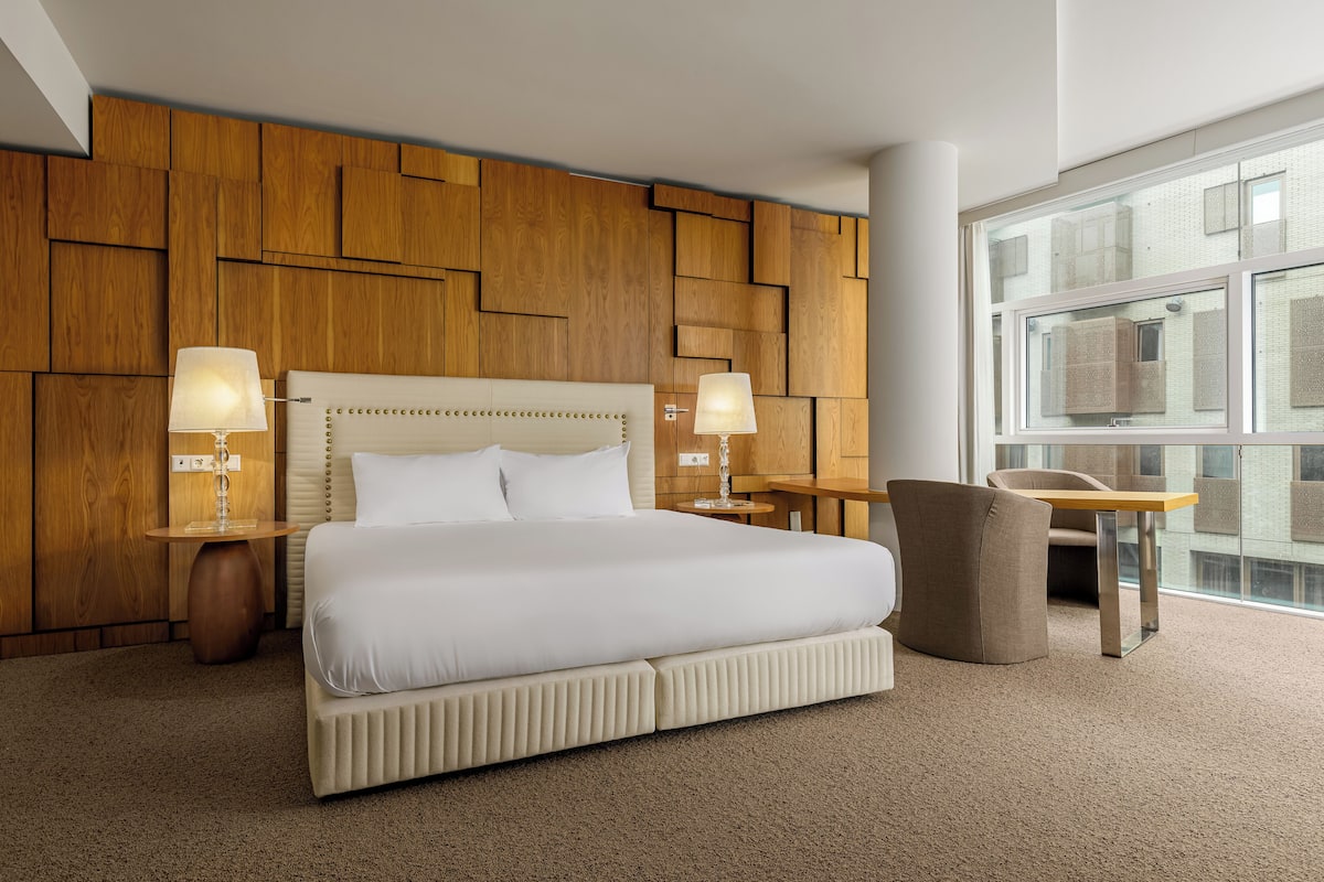 Habitación de hotel con una cama grande, dos lámparas de noche, una pared decorativa de madera, una ventana y una pequeña mesa con una silla.